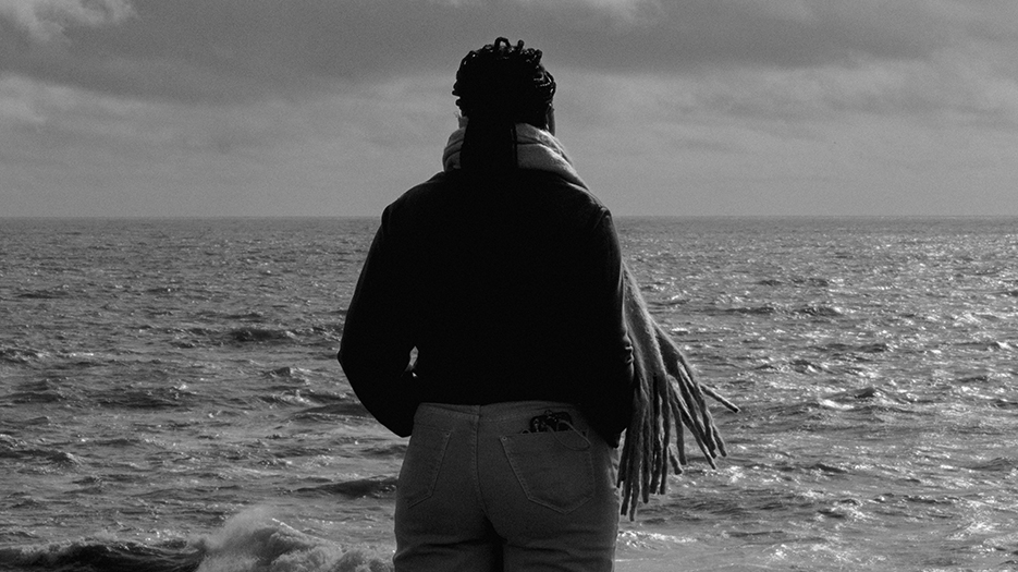 Photo en noir et blanc d'une jeune femme noire, de dos, qui regarde l'océan. Elle a les cheveux tressés réunis en queue de cheval, ses mains dans les poches de son jean, son écharpe au vent et son smartphone qui dépasse de la poche arrière de son jean.