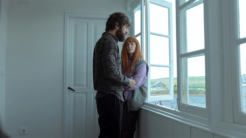 Les personnages principaux, Alex et Judy, près d'une fenêtre ouverte. Alex fume une cigarette, l'air concerné et fatigué. Judy, une femme aux longs cheveux roux, est à l'écoute.