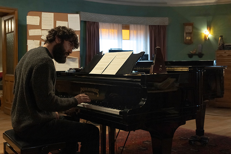 Le protagoniste, Alex, assis au piano. Alex est un homme grand, de carrure doucement musclée. Il est brun, les cheveux légèrement bouclés, et il a une grosse barbe brune. Le piano est à queue.
