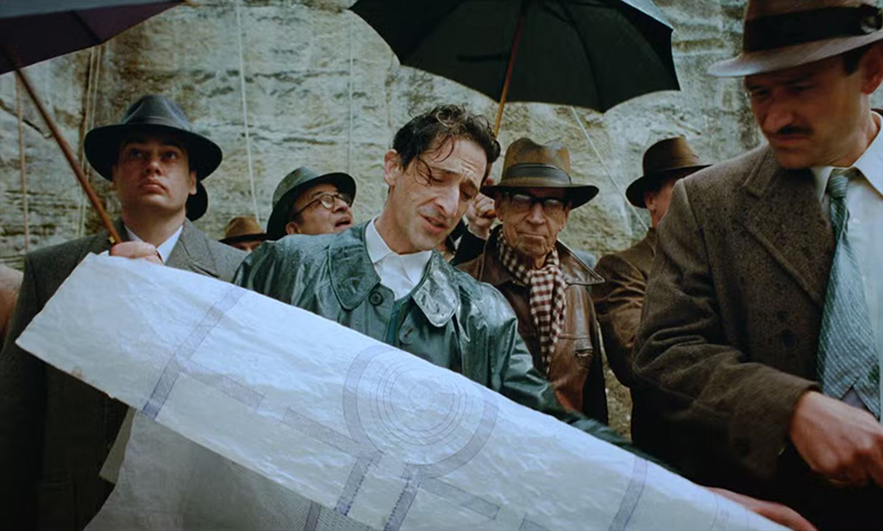 Adrien Brody dans le rôle de l'architecte. Il montre un plan à un groupe d'hommes.