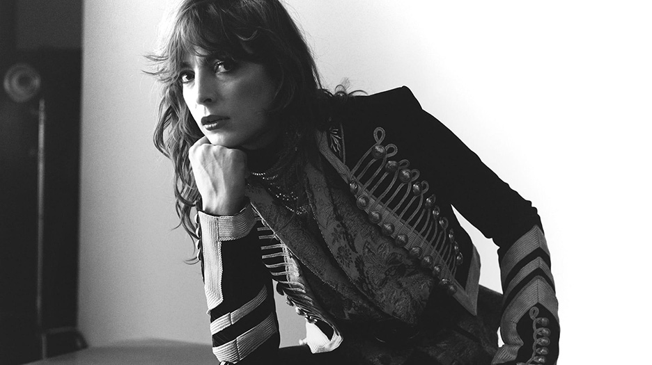 Photo en noir et blanc de Flora Fishbach par Denis Boulze pour Les Inrocks. Elle repose sa tête sur son poing, le coude sur la cuisse. Elle porte une veste de Monsieur Loyal et porte ses cheveux longs détachés.