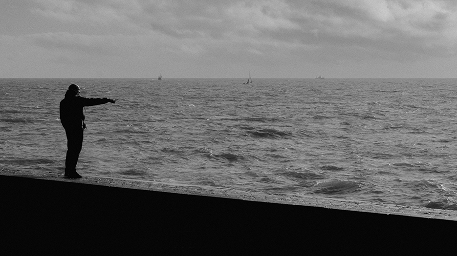 Photo en noir et blanc d'un homme debout sur la jetée, l'océan en arrière plan. L'homme pointe quelque chose du doigt mais on ne voit pas quoi.