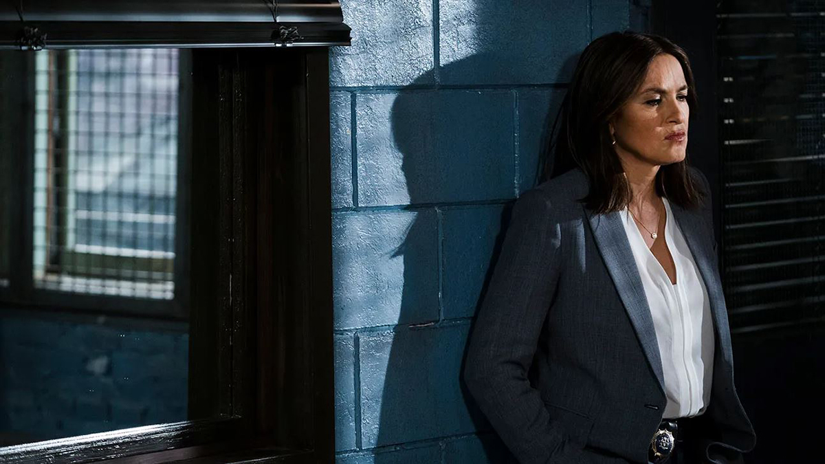 Olivia Benson, personnage principal de New York Unité Spécial. C'est ici une femme dans la fleur de l'âge, elle est adossée à un mur de grosses briques dans une salle d'interrogatoire. Elle porte une veste de blazer sur un t-shirt blanc et semble contrariée, ou en colère.