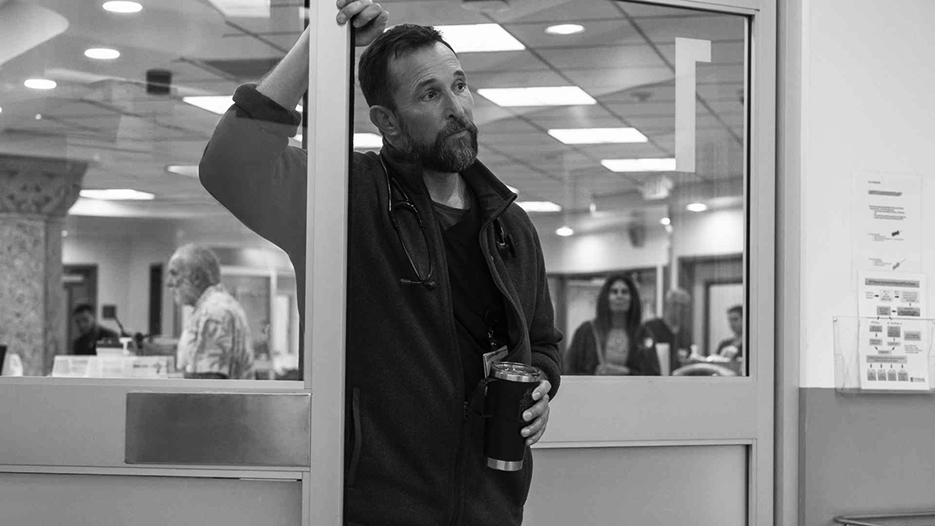 Noah Wyle dans The Pitt. C'est un homme cinquantenaire blanc barbu en tenue de soignant qui s'appuie contre une porte vitrée. Il tient un mug de café dans son autre main et il a le regard dans le vague.