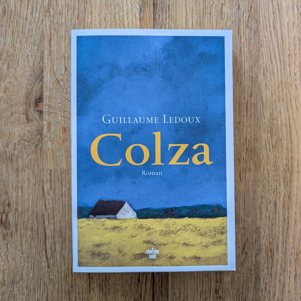 La couverture représente un champ de colza avec une maison au loin et un ciel bleu.