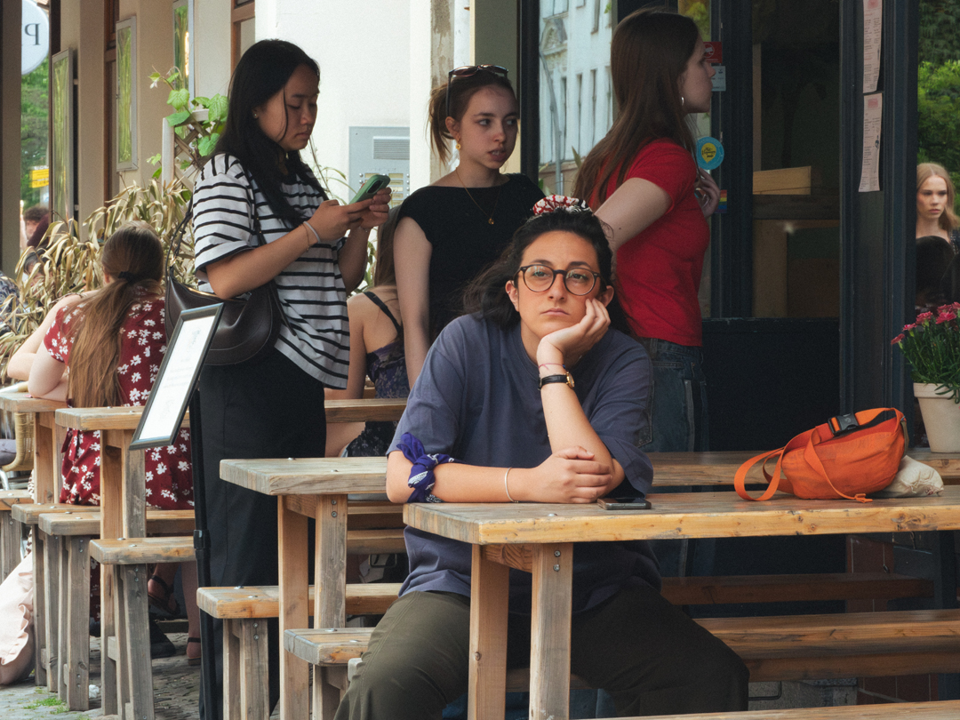 À la terrasse d'un café, dans la rue, une jeune femme est assise, un peu affalée, les jambes écartées et sa main gauche soutenant sa tête via son menton. Elle regarde dans le vide. Derrière elle, trois jeunes filles entrent dans le café et contrastent avec la première femme. La jeune femme est vêtue à la cool, un peu normcore, tandis que les trois jeunes filles sont plus mode, plus dans le coup.