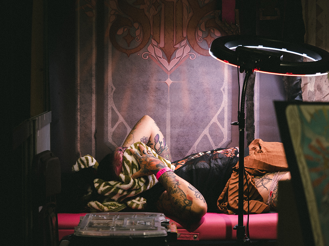 Une femme est allongée sur la table d'un tatoueur, le visage couvert par son sweat zébré vert et blanc. Elle a l'air de souffrir. Cependant on ne voit pas le tatoueur. Une lampe d'appoint est braquée sur cette femme, provoquant des lumières rosées, notamment dues au décors de l'arrière-plan.