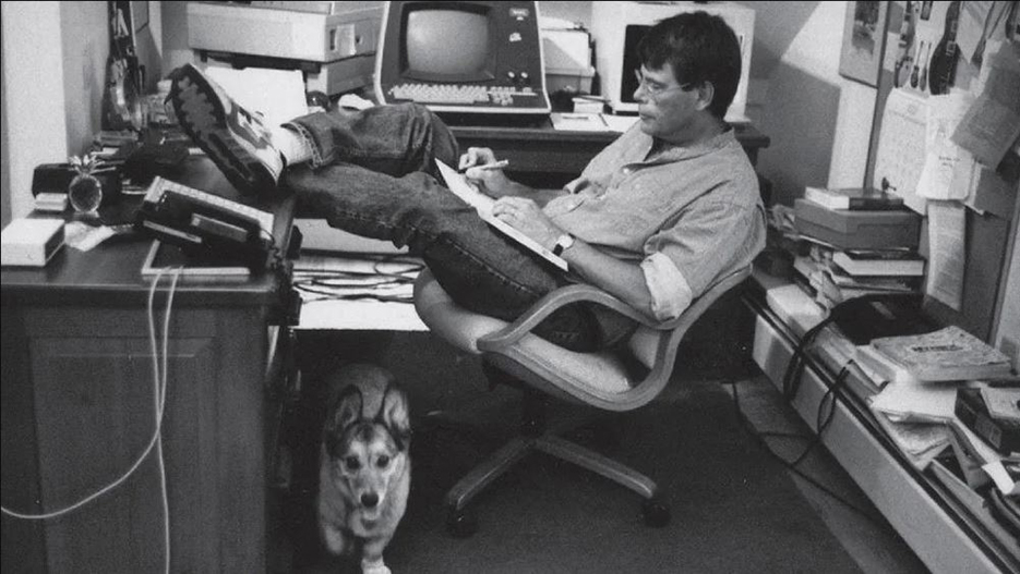 Vieille photo en noir et blanc de Stephen King en train d'écrire à la main dans un cahier, les pieds posés sur son bureau et son Corgi sous les jambes.