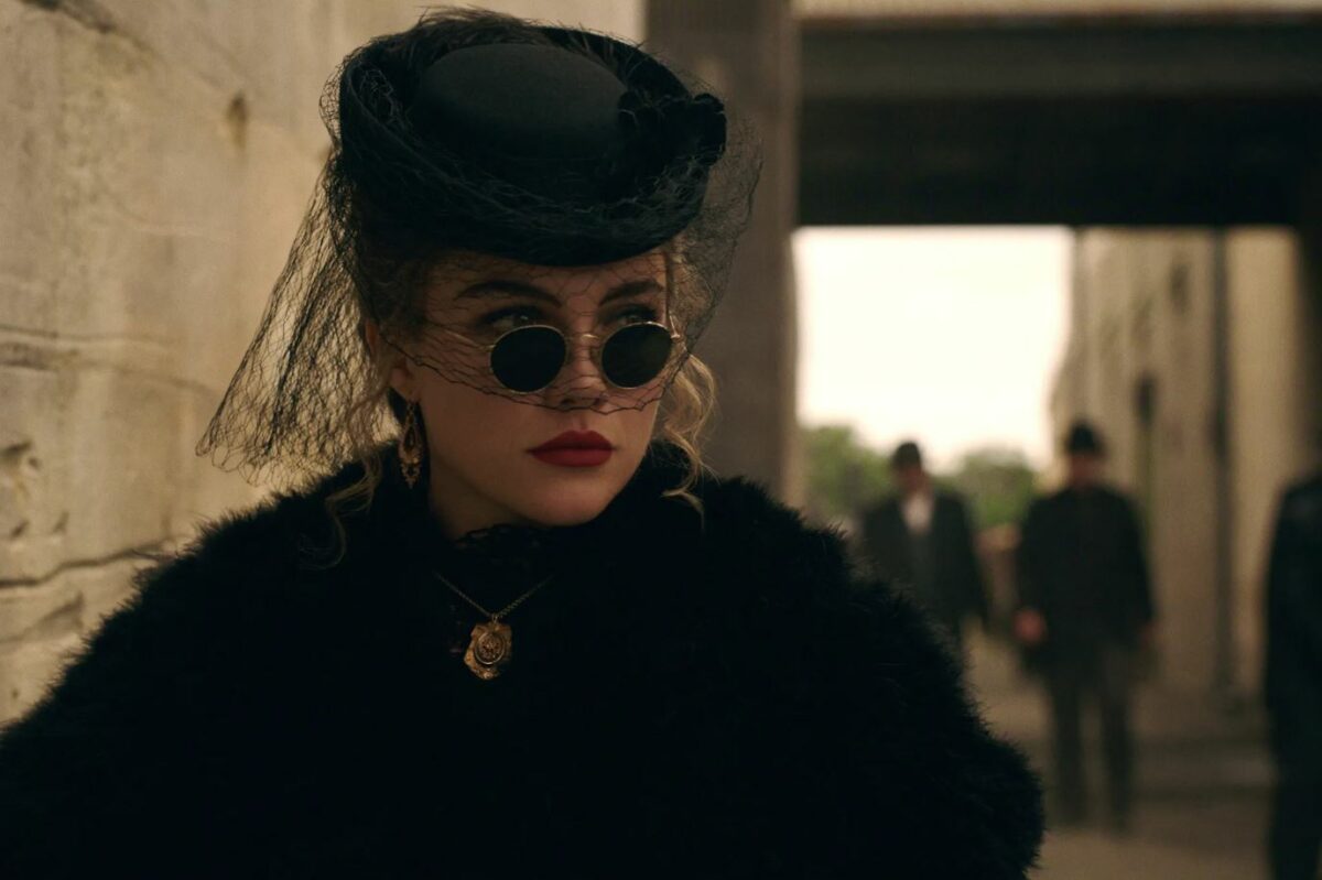 Florence Pugh dans le rôle de Cathy dans East of Eden. Habillée de noir, elle porte un chapeau à voilette et des lunettes de soleil à la mode du début du XXe siècle.