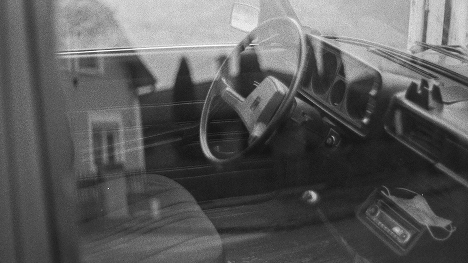 L'habitacle d'une voiture des années 80 ou 90 vu depuis derrière la vitre, créant un effet de double-exposition.