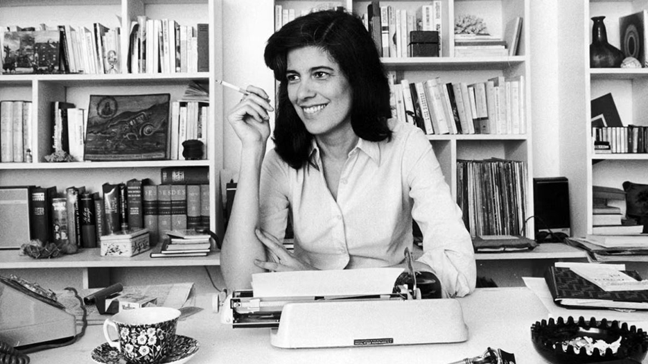 Susan Sontag, souriante, derrière sa machine à écrire, avec une cigarette à la main et une tasse de thé à proximité.