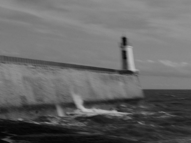 Photo en noir et blanc et floue d'un phare au bout d'une jetée. Les vagues explosent contre la digue.
